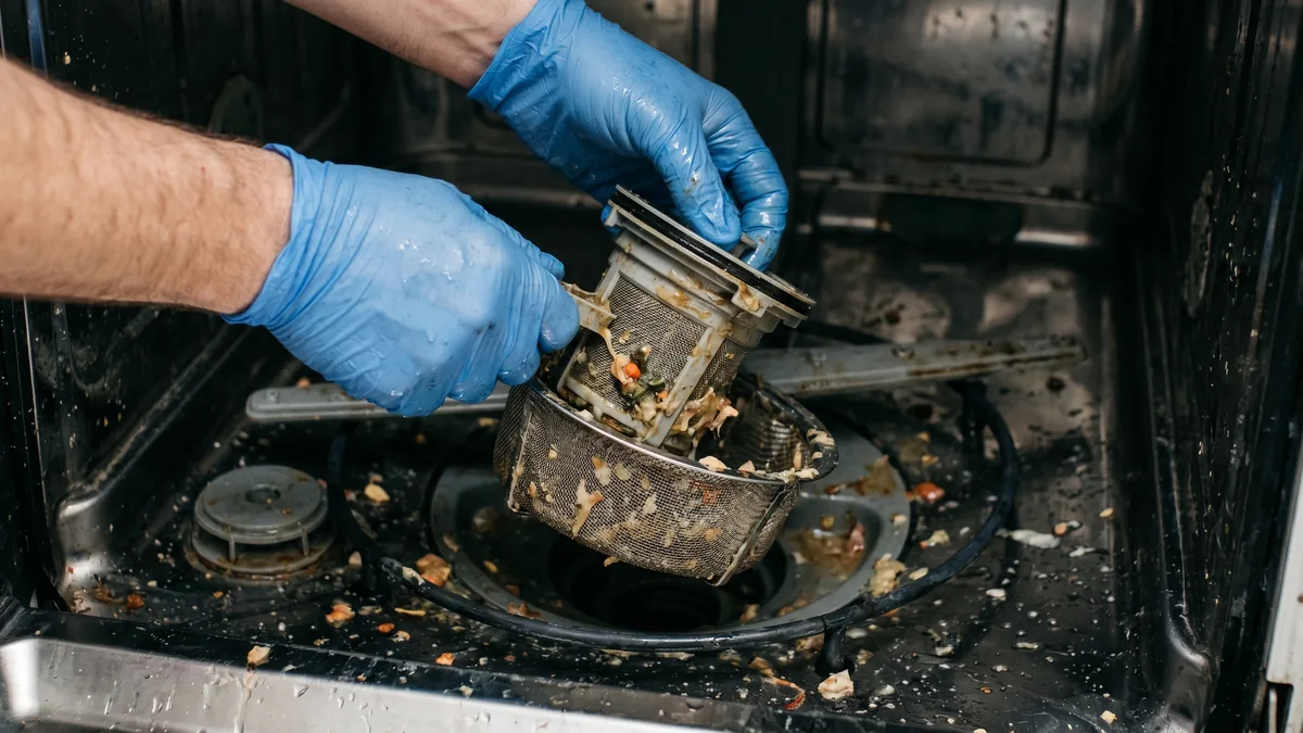 Gloved hands removing a dishwasher drain basket with visible food debris